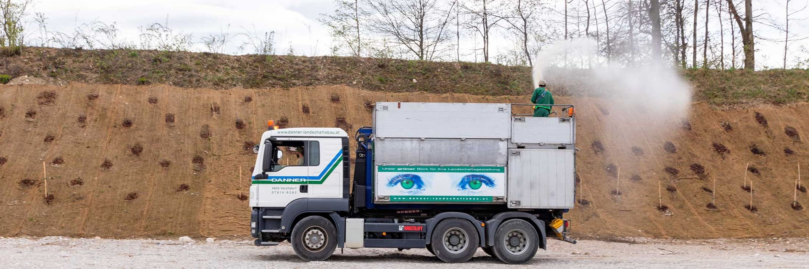 Erosionsschutz durch Kokosgewebe mit Aufspritzbegrünung, die auf dem Anhänger eines Lkw von einem Arbeiter am Hang aufgetragen wird.